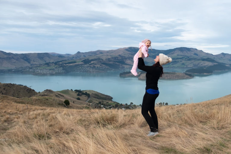 Jennifer from Backyard Travel Family lifts baby Emilia, while overlooking Banks Peninsula from the Rapaki Track Walk, Christchurch, Canterbury