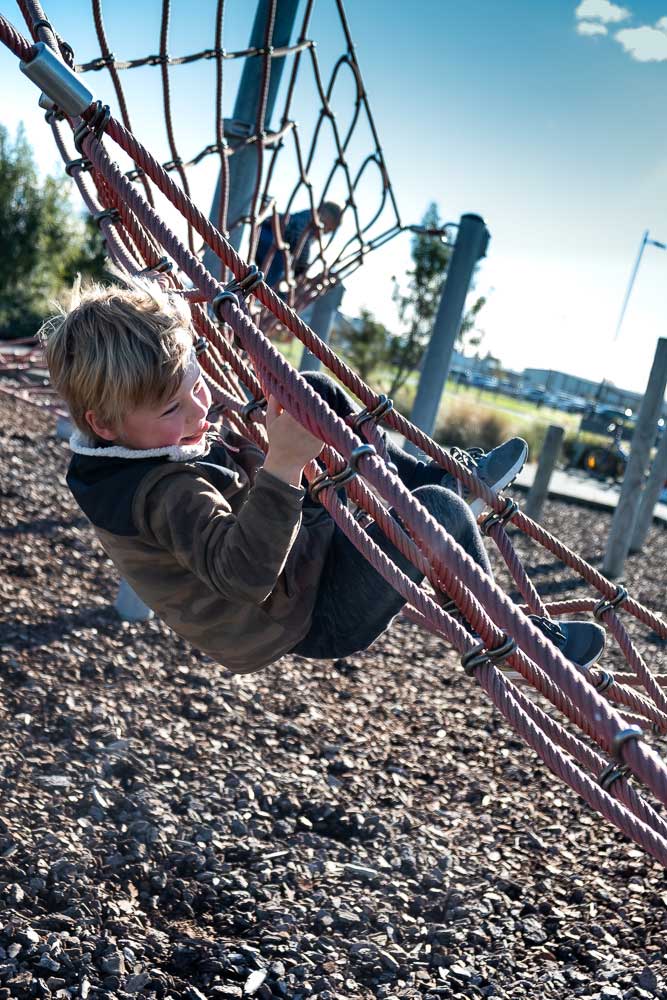 Nathan from Backyard Travel Family clings onto the rope climbing structure at Neil Graham Park Playground, Halswell, Christchurch, New Zealand