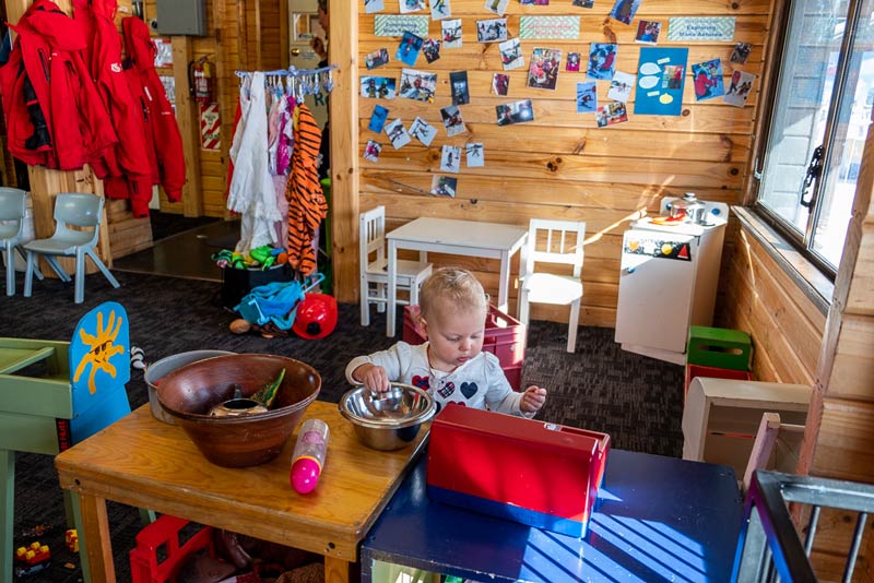 Wide angle of Skiwiland Preschool, inside Emilia plays with the cash register while her brother skis outside.