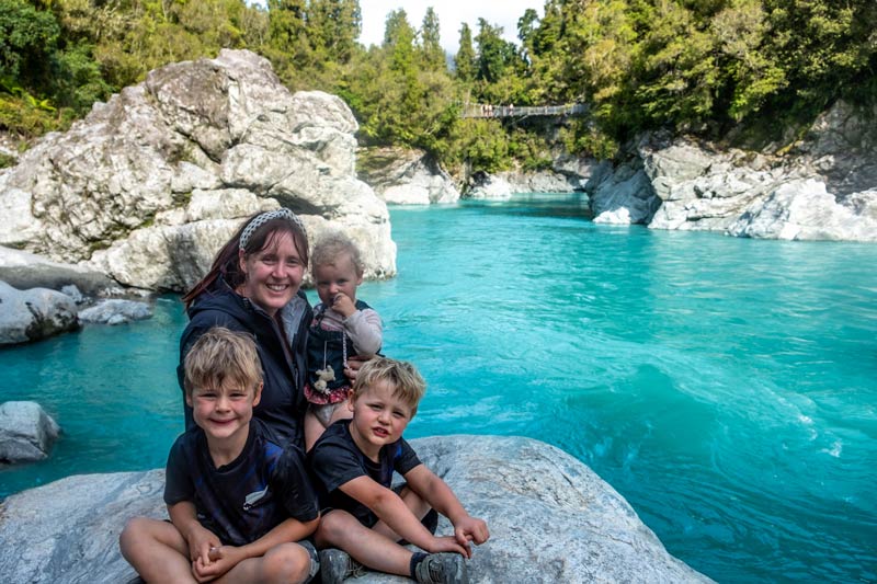 Backyard Travel Family sit on a rock at the Hokitika Gorge, with the swing bridge in the background. The crazy blue water shows that this really is one of the best things to do in Hokitka