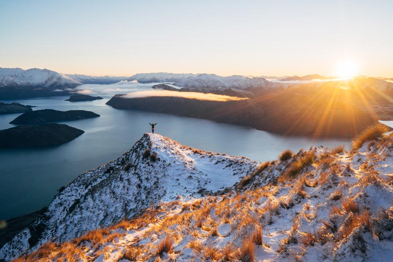 A person stands on the ledge of the Roys Peak Track at sunrise, sun peaking over the mountains, arms up in awe as they look at the mountains and Lake Wanaka below