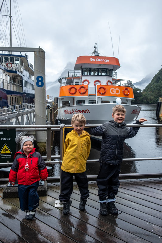 Backyard Travel Family kids stand in front of the Go Orange Milford Haven boat before a Milford Sound Cruise