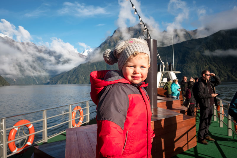 2 year old Emilia, standing on the top deck of Go Orange Milford Sound Cruise boat, looking at the amazing waterfalls