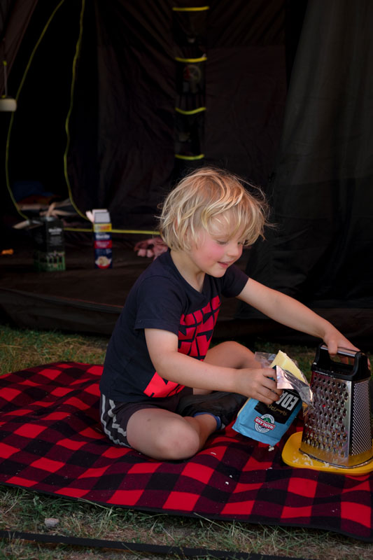 Kipton from Backyard Travel Family grates cheese for dinner while sitting on a plaid picnic mat