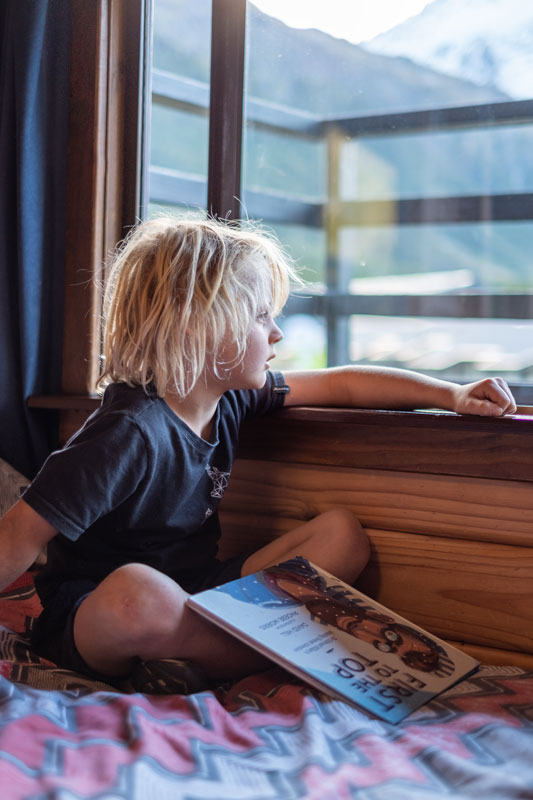 Young boy reading a book, looking out the window at the mountains at YHA Aoraki Mt Cook