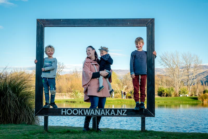 Backyard Travel Family stand inside a wooden frame, in front of a manmade lake with salmon at Hook Wanaka NZ