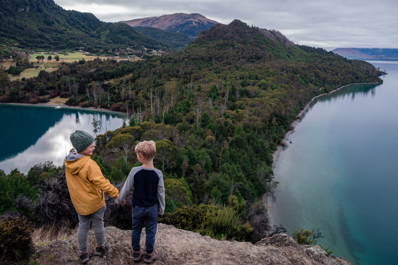 Backyard Travel Family boys hold hands while they look lovingly at each other as they look over Lake Wakatipu and Bobs Cove in Queenstown, New Zealand