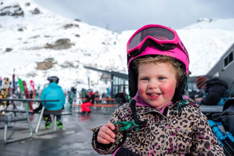 Emilia from Backyard Travel Family, wearing a pink ski hement and ski jacket, sits on a picnic table at the Remarkables ski field Queenstown NZ