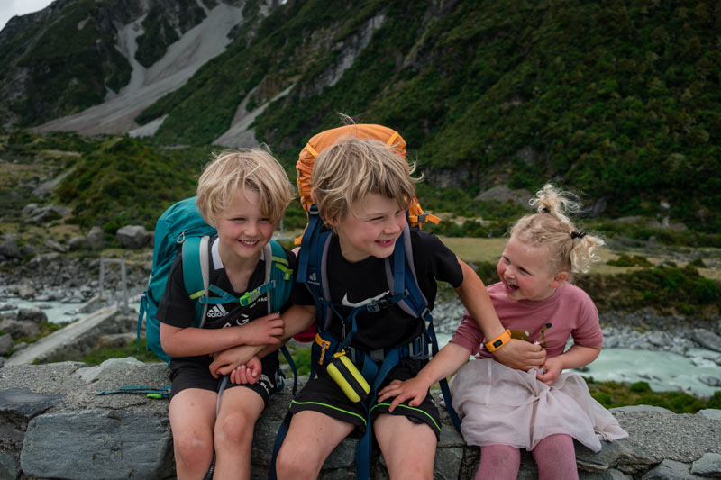 3 kids from Backyard Travel Family sit on rocks with the first swing bridge of the Hooker Valley Track in behind. This is the start of the Hooker Hut track