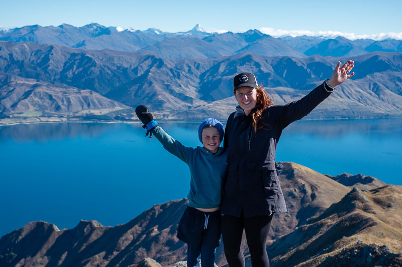 Nathan and Jen from Backyard Travel Family lift their arms in delight as they reach the summit of Breast Hill, the incredible blue lake Hawea in the background along with the snowy tip of Mt Aspiring