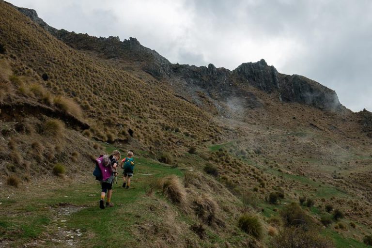 2 children walk up a grassy 4wd section of the Isthmus Peak track, mountains towering in behind