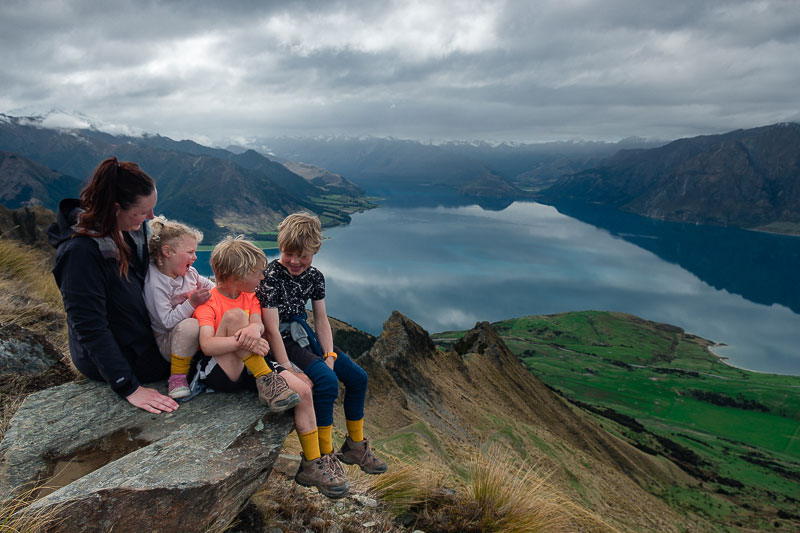 Family sit on a rock overlooking Lake Hawea on the Ishtmus Peak Hike, a doable but challenging walk for kids