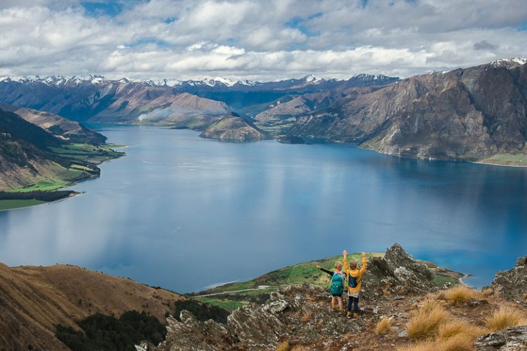 Incredible viewpoint overlooking the blue Lake Hawea, with snow capped mountains in the distance. 2 children have their hands in the air, stoked to have made it so far on the Isthmus Peak track