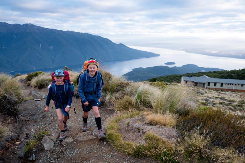 2 young boys walk up the hill on day 2 of the Kepler Track with kids, the wooden lodge of Luxmore Hut and Lake Te Anau in view, showing off how high the boys have climbed