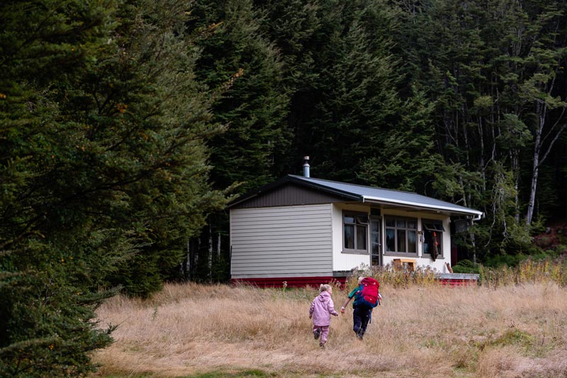 View of the white wooden Kiwi Burn Hut building nestled at the edge of the forest as two children run towards the hut